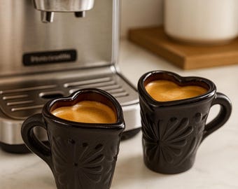 May include: Two black heart-shaped coffee mugs filled with espresso. The mugs have a floral design and are placed in front of a stainless steel espresso machine. A white and brown container is in the background.