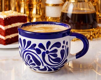 Op de afbeelding: Een wit en blauw keramische koffiemok gevuld met een lichtbruine drank. De mok heeft een blauw oor en is versierd met een blauw bloemmotief. Een plakje red velvet cake ligt links op een bord.