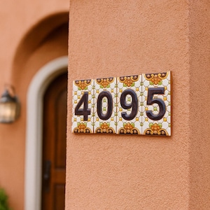 Pode incluir: Azulejos decorativos com o número da casa 4095. Os números castanhos escuros destacam-se num fundo branco com um desenho floral em amarelo, laranja e verde. Os azulejos estão montados numa parede texturizada cor de pêssego.