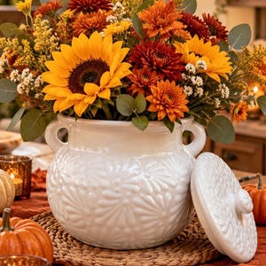 Puede incluir: Una olla de cerámica blanca con tapa, dos asas laterales y un patrón floral texturizado, llena con un ramo otoñal de girasoles amarillos, crisantemos de color rojo intenso, flores naranjas y hojas de eucalipto verdes.