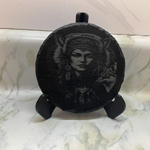 Puede incluir: Un posavasos de pizarra negra con una imagen grabada de una mujer con un lobo. La mujer lleva un tocado y plumas en el pelo. El lobo mira hacia un lado. El posavasos está sobre un soporte negro.