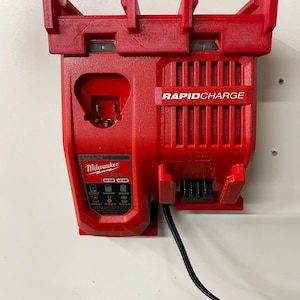 以下が含まれることがあります： 白い壁に取り付けられた赤いミルウォーキーラピッドチャージバッテリー充電器。充電器には黒い電源コードと「M12 M18」と書かれたラベルが付いています。