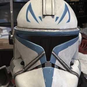 May include: A white and blue clone trooper helmet featuring a weathered finish, a central ridge, and blue angular markings above the visor. The helmet includes a side-mounted antenna, a T-shaped visor opening, and two circular vents at the chin.