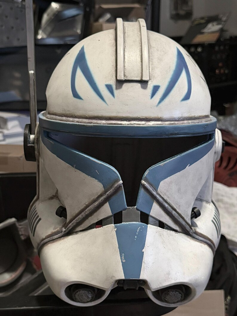 May include: A white and blue clone trooper helmet featuring a weathered finish, a central ridge, and blue angular markings above the visor. The helmet includes a side-mounted antenna, a T-shaped visor opening, and two circular vents at the chin.