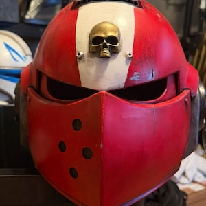 Könnte beinhalten: Ein rot-weißer Vollvisierhelm mit verwittertem Finish und einem metallisch goldfarbenen Totenkopf-Emblem in der Mitte der Stirn. Der Helm verfügt über einen horizontalen Augenschlitz und eine untere Frontplatte mit vier runden Lüftungslöchern.