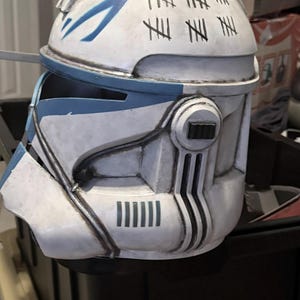 May include: A white and blue clone trooper helmet featuring a weathered finish with black tally marks painted on the top. The helmet includes a T-shaped visor, side ear caps with black rectangular details, and vertical vent markings on the lower jaw area.