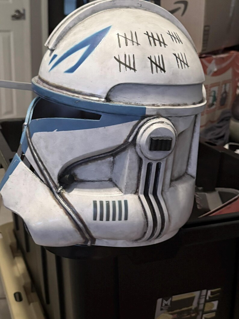 May include: A white and blue clone trooper helmet featuring a weathered finish with black tally marks painted on the top. The helmet includes a T-shaped visor, side ear caps with black rectangular details, and vertical vent markings on the lower jaw area.