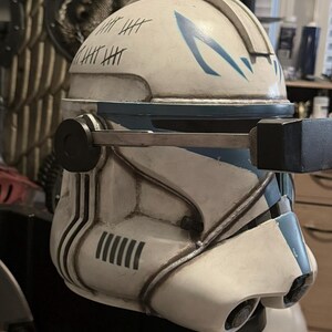 May include: A white and blue clone trooper helmet featuring a weathered finish, black tally marks on the side, and a mechanical rangefinder attachment extending from the side. The helmet has a blue chevron design on the forehead and dark accents.