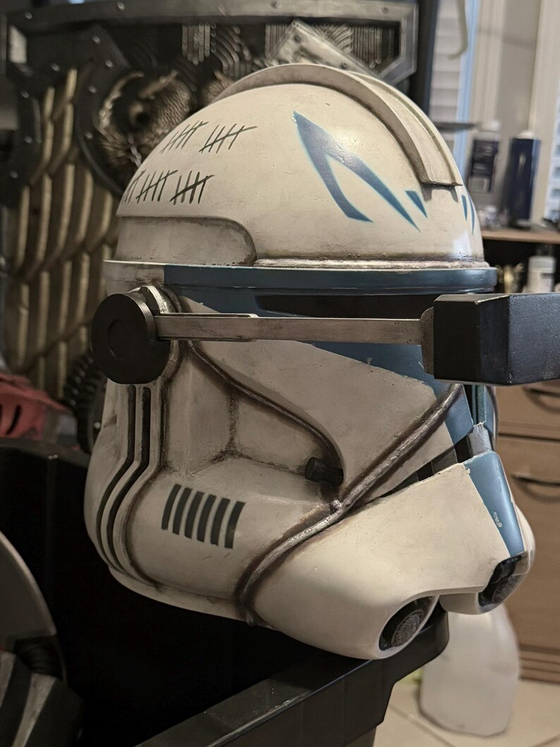 May include: A white and blue clone trooper helmet featuring a weathered finish, black tally marks on the side, and a mechanical rangefinder attachment extending from the side. The helmet has a blue chevron design on the forehead and dark accents.