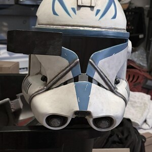 May include: A white and blue clone trooper helmet featuring a weathered finish, a black rectangular rangefinder attached to the right side, a T-shaped visor, and two circular ventilation ports located on the lower front section of the faceplate.
