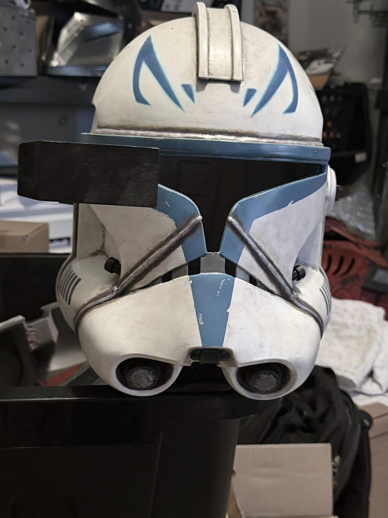 May include: A white and blue clone trooper helmet featuring a weathered finish, a black rectangular rangefinder attached to the right side, a T-shaped visor, and two circular ventilation ports located on the lower front section of the faceplate.