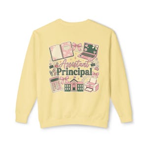 May include: Pale yellow sweatshirt with a colorful graphic design. The design includes a notebook, laptop, calculator, printer, schoolhouse, and bows. The words "Assistant Principal" are in the center of the design.