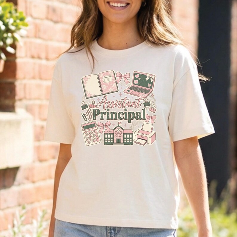 May include: Cream-colored t-shirt with a colorful graphic design. The design includes a notebook, laptop, calculator, schoolhouse, and printer, with the words "Assistant Principal" in the center. Pink bows and accents are incorporated throughout the design.