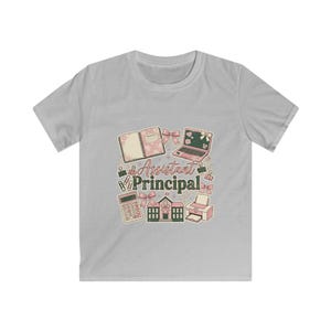 May include: A light gray t-shirt featuring a graphic design with school-related elements. The design includes a notebook, laptop, calculator, school building, and printer, with the text "Assistant Principal".
