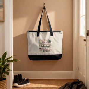May include: A canvas tote bag with black handles and a black bottom panel. The bag features a design with the words "Assistant Principal" and illustrations of books, a laptop, and other school-related items. The bag is hanging on a wall.