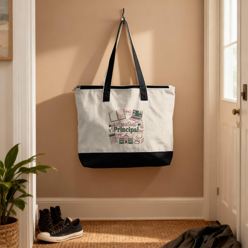 May include: A canvas tote bag with black handles and a black bottom panel. The bag features a design with the words "Assistant Principal" and illustrations of books, a laptop, and other school-related items. The bag is hanging on a wall.