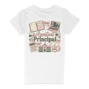 May include: White t-shirt with a colorful graphic design. The design includes a notebook, laptop, calculator, school building, and printer, with the words "Assistant Principal" in the center. Pink and green accents.