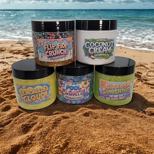 May include: Five jars of slime with black lids are displayed on a sandy beach with the ocean in the background. The jars are labeled "Flip Flop Crunch," "Coconut Cream," "Lemonade Cloud," "Pool Floatle," and "Citrus Sunshine."