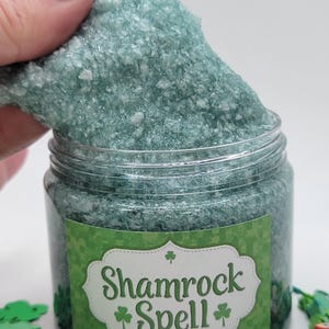May include: A clear jar filled with green, crystalline bath salts. The jar has a green and white label that reads "Shamrock Spell" with shamrock graphics. A hand is holding a portion of the bath salts above the jar.