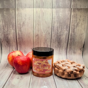 May include: A jar with a black lid and a label that reads "Apple Pie Pop" next to three red apples and a brown pie-shaped object. The label features a watercolor illustration of an apple pie.