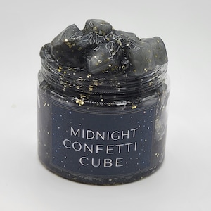 May include: A clear jar filled with black slime and gold glitter. The jar has a dark blue label with the words "MIDNIGHT CONFETTI CUBE" in white. The slime is piled on top of the jar, giving it a textured appearance.