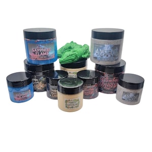 May include: An assortment of small, round jars with black lids, each labeled with a unique name. The jars feature various colors and designs, including "Sapphire Flame," "Ancient Smoke," and "Forest Hatchling." A green dragon figurine sits atop one of the jars.