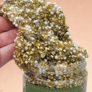 May include: A clear jar filled with slime, featuring a mix of gold and white beads. The slime is a textured, viscous material, with a portion spilling over the jar's rim. The background is a solid peach color.