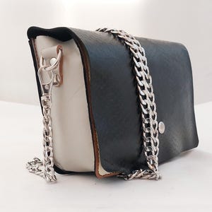 May include: A structured handbag featuring a black textured flap and white side panels. The bag includes a silver-toned metal chain strap attached with lobster claw clasps and a single silver snap button closure on the front flap.