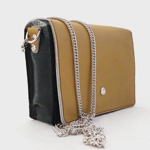 Puede incluir: Un bolso de hombro rectangular con una solapa frontal de color tostado, un cierre de presión plateado y un panel lateral negro. El bolso incluye una correa de cadena de metal plateado sujeta a los lados con herrajes de color plata.