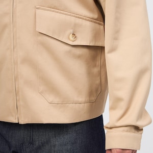 May include: A tan jacket featuring a buttoned pocket and a zip. The jacket has long sleeves and is made of a smooth fabric. The bottom of the image shows dark blue denim jeans.