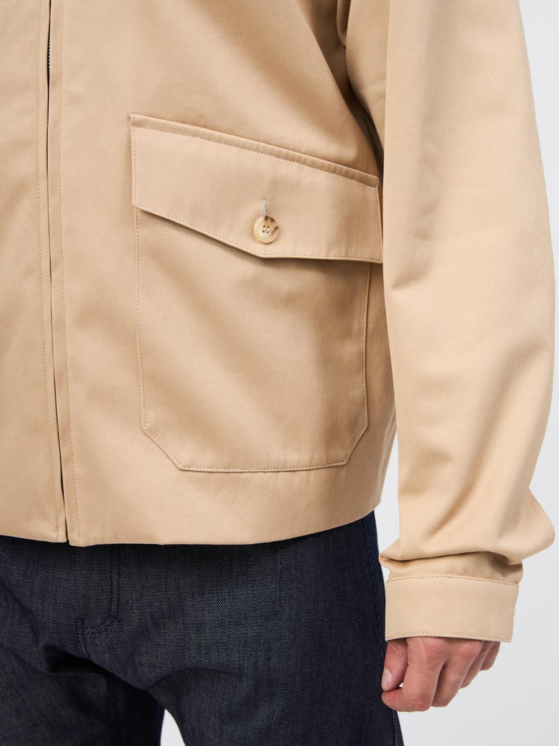 May include: A tan jacket featuring a buttoned pocket and a zip. The jacket has long sleeves and is made of a smooth fabric. The bottom of the image shows dark blue denim jeans.