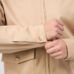 May include: A beige jacket with a zip fastening and buttoned cuffs. The jacket features two front pockets with button closures. The sleeves are being buttoned. The jacket is made of a plain, solid-coloured fabric.