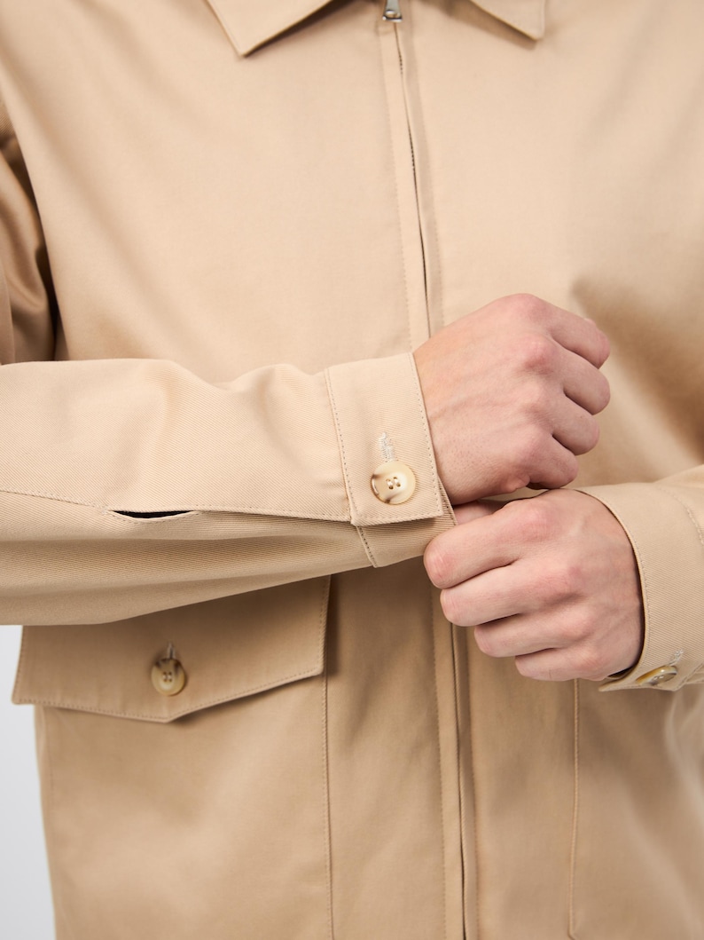 May include: A beige jacket with a zip fastening and buttoned cuffs. The jacket features two front pockets with button closures. The sleeves are being buttoned. The jacket is made of a plain, solid-coloured fabric.