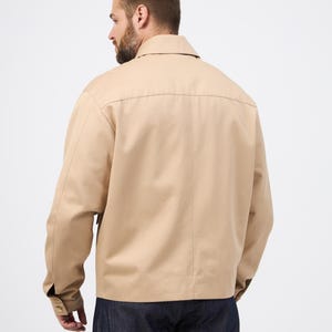 May include: A beige jacket featuring a collared neckline and buttoned cuffs. The jacket is made of a smooth material and has a simple, classic design. The model is wearing dark blue jeans. A versatile piece of outerwear.