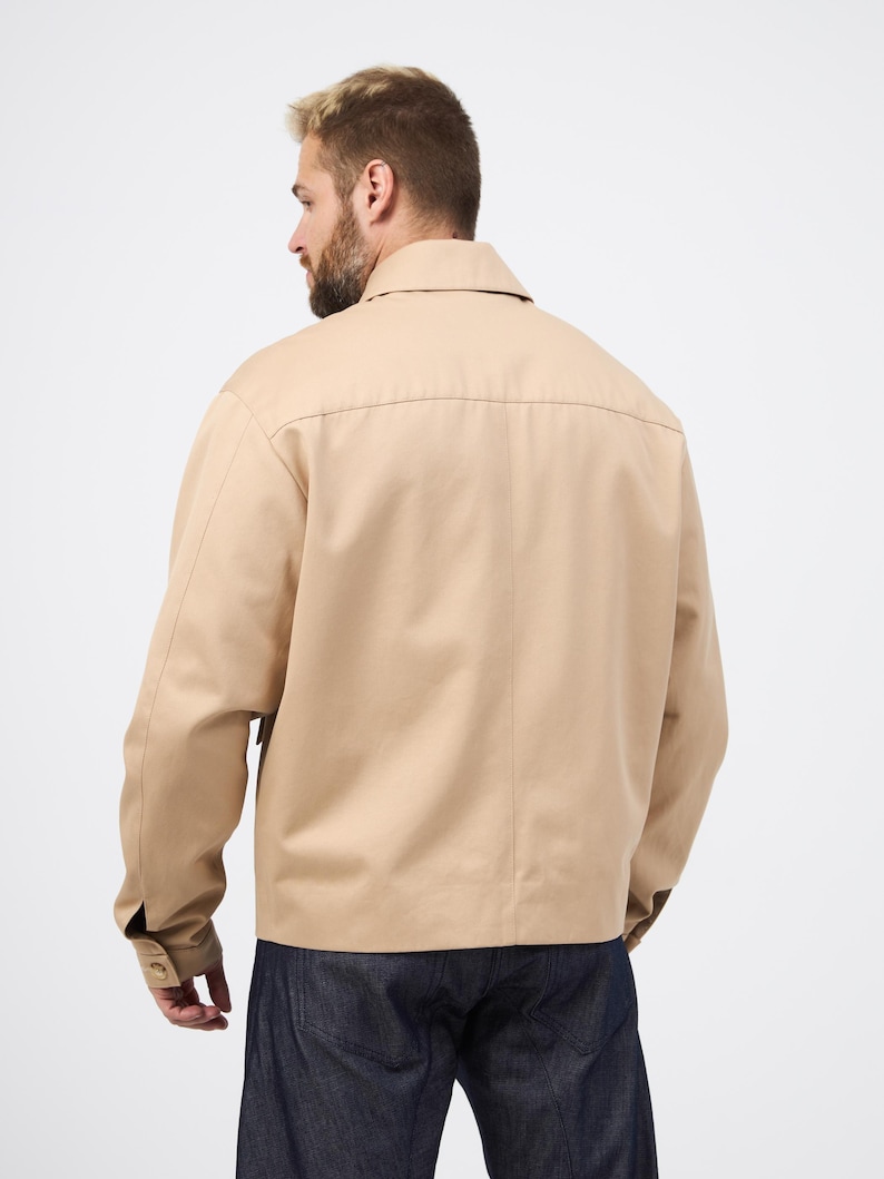 May include: A beige jacket featuring a collared neckline and buttoned cuffs. The jacket is made of a smooth material and has a simple, classic design. The model is wearing dark blue jeans. A versatile piece of outerwear.