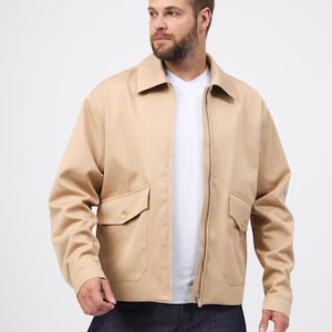 May include: A beige jacket with a zip fastening and two front pockets. The jacket features a collared neckline and long sleeves. The person in the image is wearing a white t-shirt and dark blue jeans.