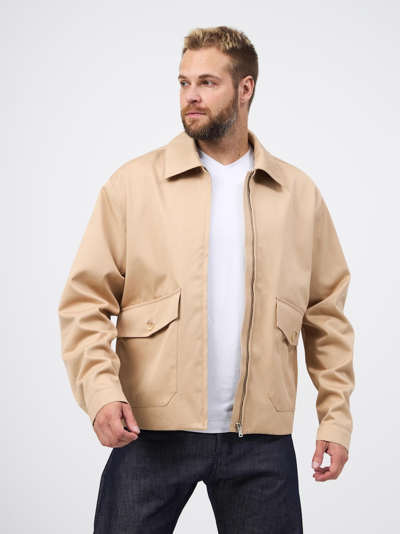 May include: A beige jacket with a zip fastening and two front pockets. The jacket features a collared neckline and long sleeves. The person in the image is wearing a white t-shirt and dark blue jeans.
