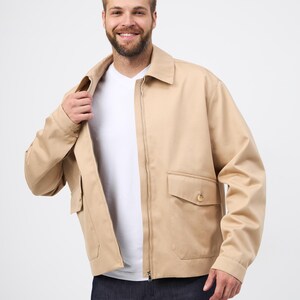 May include: A beige jacket with a zip fastening, two front pockets, and a collared neckline. The jacket is worn over a white t-shirt and paired with dark blue jeans. The jacket's design is simple and classic.
