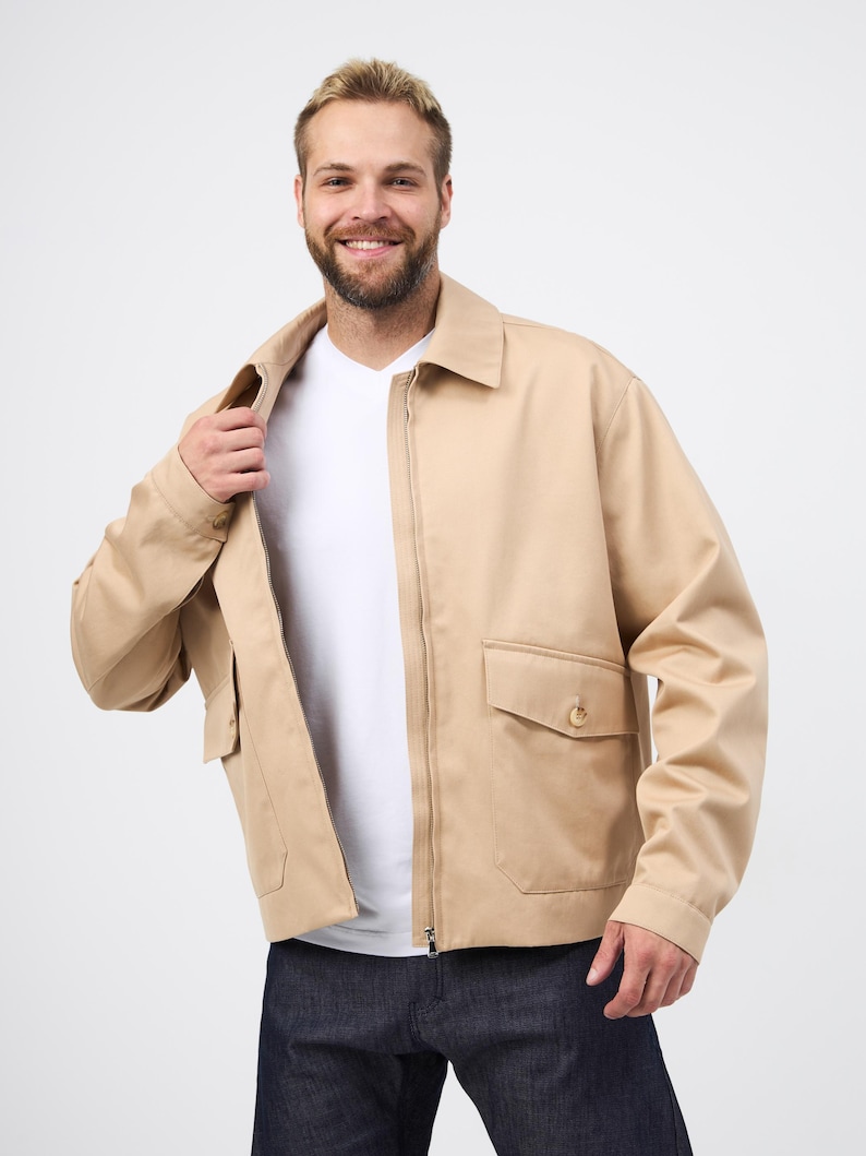 May include: A beige jacket with a zip fastening, two front pockets, and a collared neckline. The jacket is worn over a white t-shirt and paired with dark blue jeans. The jacket's design is simple and classic.