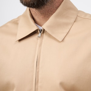 May include: A close-up shot of a tan jacket featuring a silver zip closure. The jacket has a classic collar and a minimalist design. The fabric looks to be a smooth, woven textile. The jacket's neutral tone makes it versatile for different ensembles.