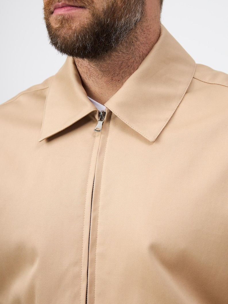 May include: A close-up shot of a tan jacket featuring a silver zip closure. The jacket has a classic collar and a minimalist design. The fabric looks to be a smooth, woven textile. The jacket's neutral tone makes it versatile for different ensembles.