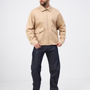 May include: A beige zip-up jacket with two front pockets and dark blue denim jeans. The jacket has a collared neckline. The jeans are straight-legged. White trainers complete the outfit. The person is standing in front of a white background.