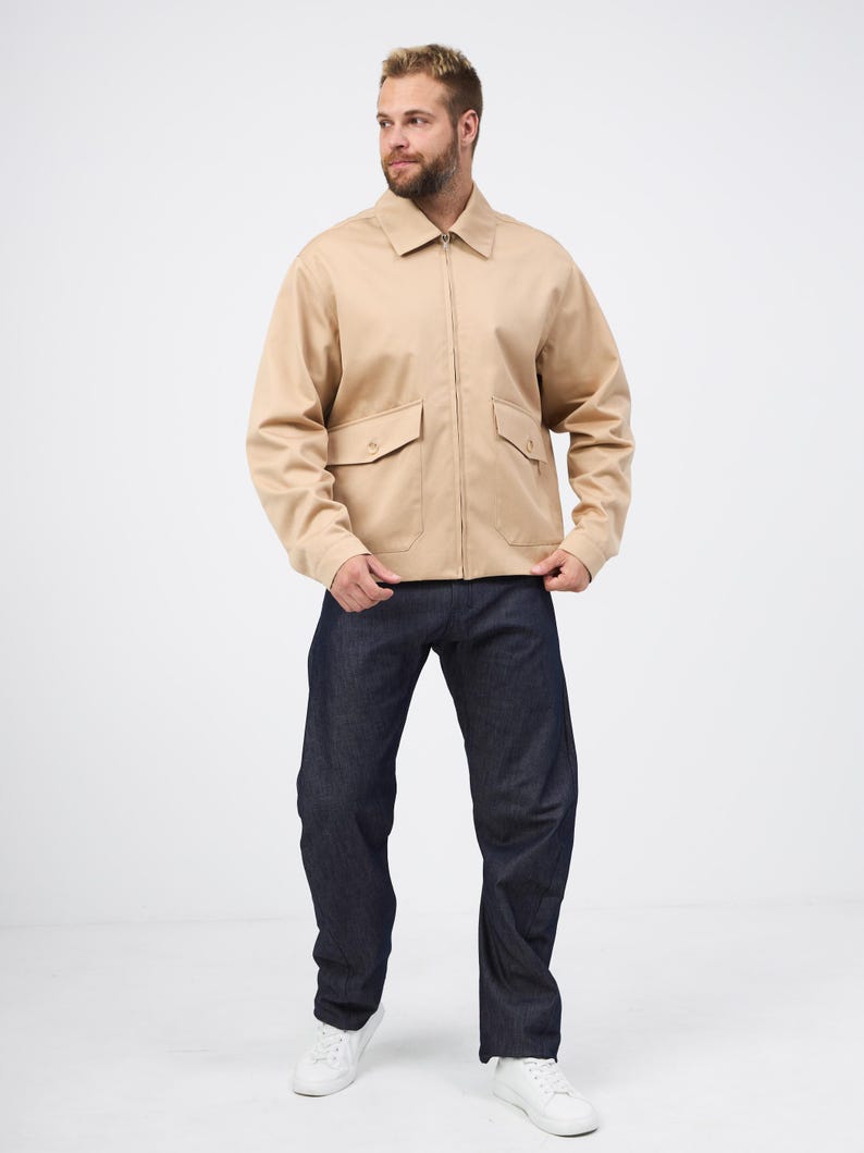 May include: A beige zip-up jacket with two front pockets and dark blue denim jeans. The jacket has a collared neckline. The jeans are straight-legged. White trainers complete the outfit. The person is standing in front of a white background.