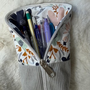 Peut inclure: Une trousse à crayons verticale avec une base en velours côtelé gris et une partie supérieure en tissu blanc ornée d'un motif floral bleu marine, orange et vert clair. La trousse est ouverte, révélant plusieurs stylos et crayons, avec une fermeture éclair argentée.
