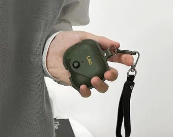 May include: A green leather case with a gold inscription that reads "Leo" and a black strap. The case has a snap closure and a metal ring for attaching a keychain.