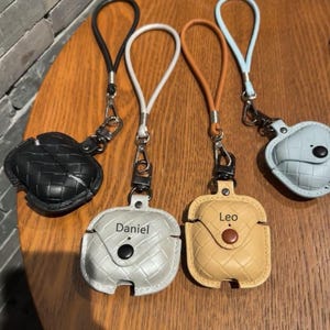 Könnte beinhalten: Drei Airpod-Hüllen mit personalisierten Namen, Daniel, Leo und einem nicht lesbaren Namen, werden auf einer Holzoberfläche gezeigt. Jede Hülle hat eine andere Farbe und ist an einem Lanyard befestigt.