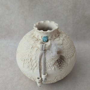 May include: A cream-colored, textured ceramic vase with a round body and a ruffled rim. It features a turquoise stone, gray suede ties, and decorative feathers. The vase has a rustic, handcrafted appearance.