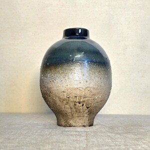 May include: A handcrafted ceramic vase with a rounded body and a narrow neck. The vase features a gradient of colors, transitioning from a dark blue top to a textured, speckled beige base. The vase is approximately 8 inches tall.