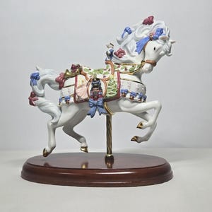 May include: A white carousel horse figurine with a detailed saddle adorned with blue bows, red accents, and gold trim. The horse stands on a wooden base, supported by a gold-colored pole. The figurine has a unicorn horn.