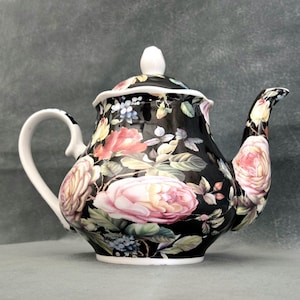 May include: Black ceramic teapot with a floral design featuring pink, yellow, and red roses. The teapot has a white handle, spout, and lid with a white finial. A decorative piece for serving tea.